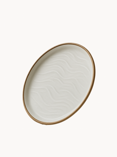 An oval ceramic platter in a matte off-white finish features a textured, wavy sand dune pattern on its interior and a natural brown rim.