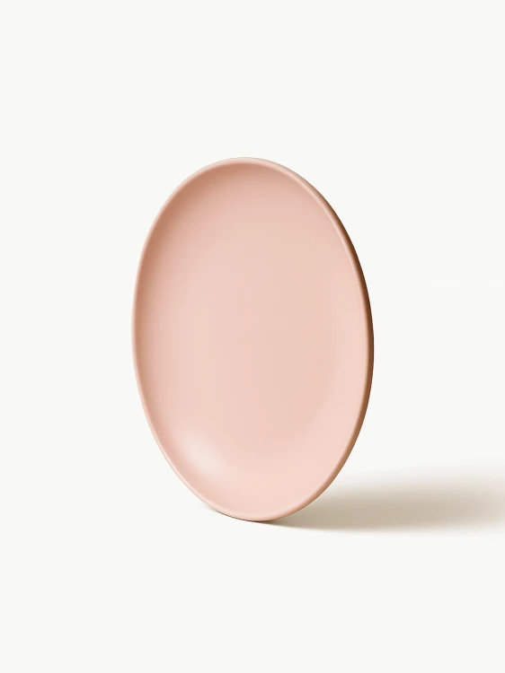 A matte, pastel pink ceramic plate. The plate is shown standing upright at a slight angle on a solid white background.