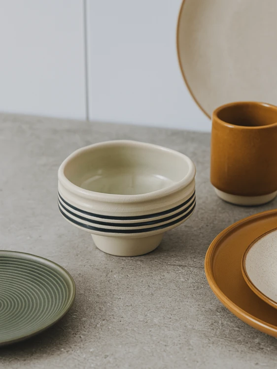 A cream-colored pedestal ceramic bowl with black horizontal stripes sits on a grey countertop. The minimalist table setting features a collection of earthy-toned plates and an ochre mug in the background.