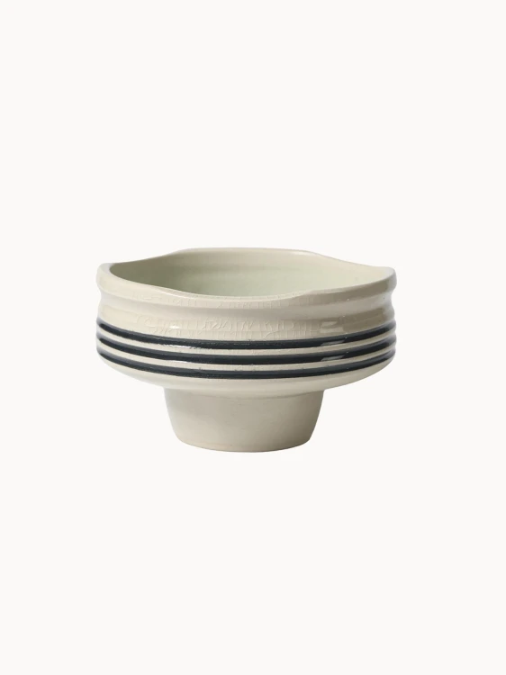 A pedestal ceramic bowl in a light cream color with a subtle crackle glaze and three horizontal black stripes around its middle.