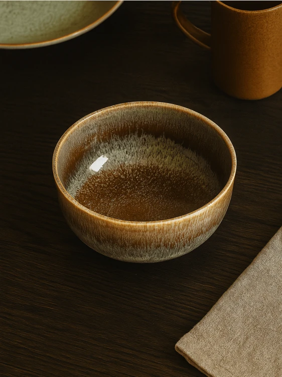 A round ceramic bowl with a dripping brown and beige glaze sits on a dark wooden table. The cozy table setting includes a linen napkin, an ochre mug, and a plate partially visible in the background.
