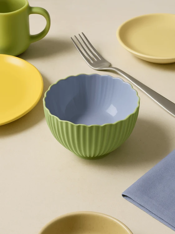 A small, two-toned ceramic bowl with a ribbed green exterior and a smooth lavender interior on a beige table. The colorful, modern table setting includes yellow plates, a green mug, a lavender napkin, and a metal fork.